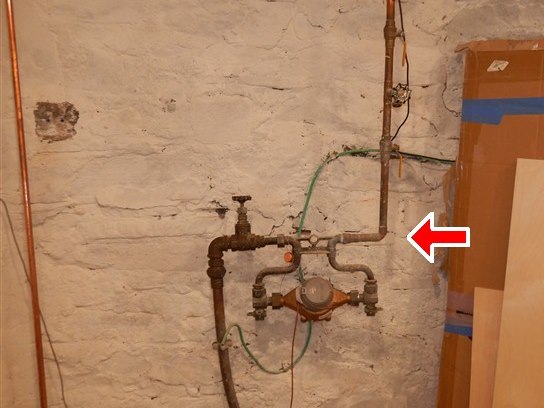 Inspection photograph of a cellar water service entry - shut-off valve, meter and copper pipework against a whitewashed stone foundation wall, with an arrow marking the item noted.